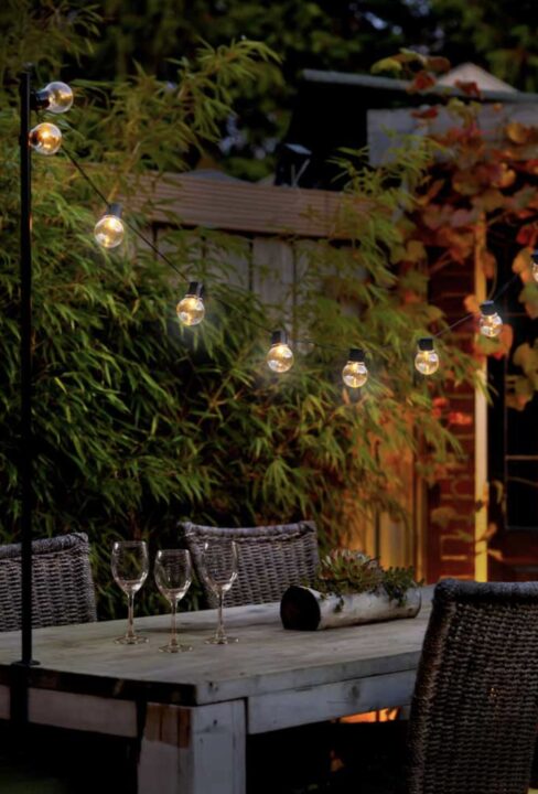 Outdoor party lights strung from table-mounted clamps, casting a warm glow over a rustic garden dining table surrounded by bamboo.