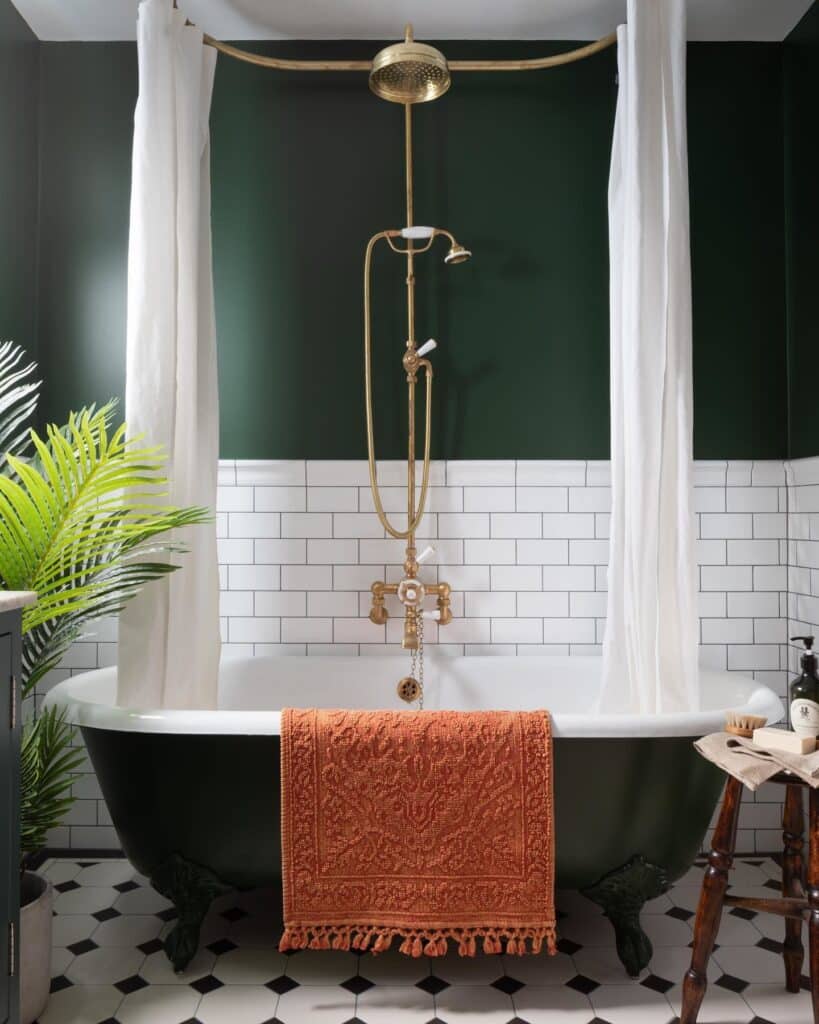 Dark green bathroom painted in Farrow and Ball Duck Green with a black clawfoot bath, white metro tiles, brass overhead shower and terracotta towel on a checker floor