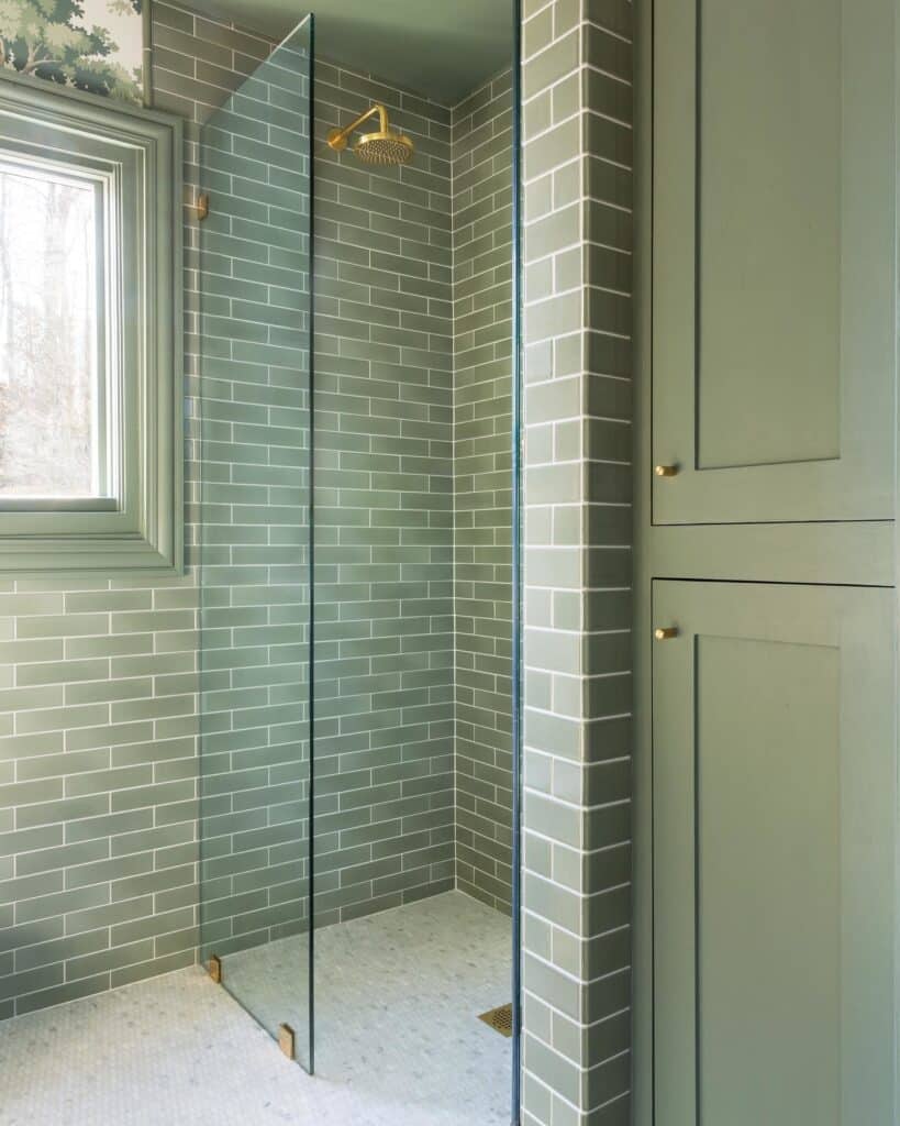 Sage green bathroom with Card Room Green subway tiles in a walk-in shower and matching painted storage cabinetry, with brass shower fittings and botanical wallpaper detail