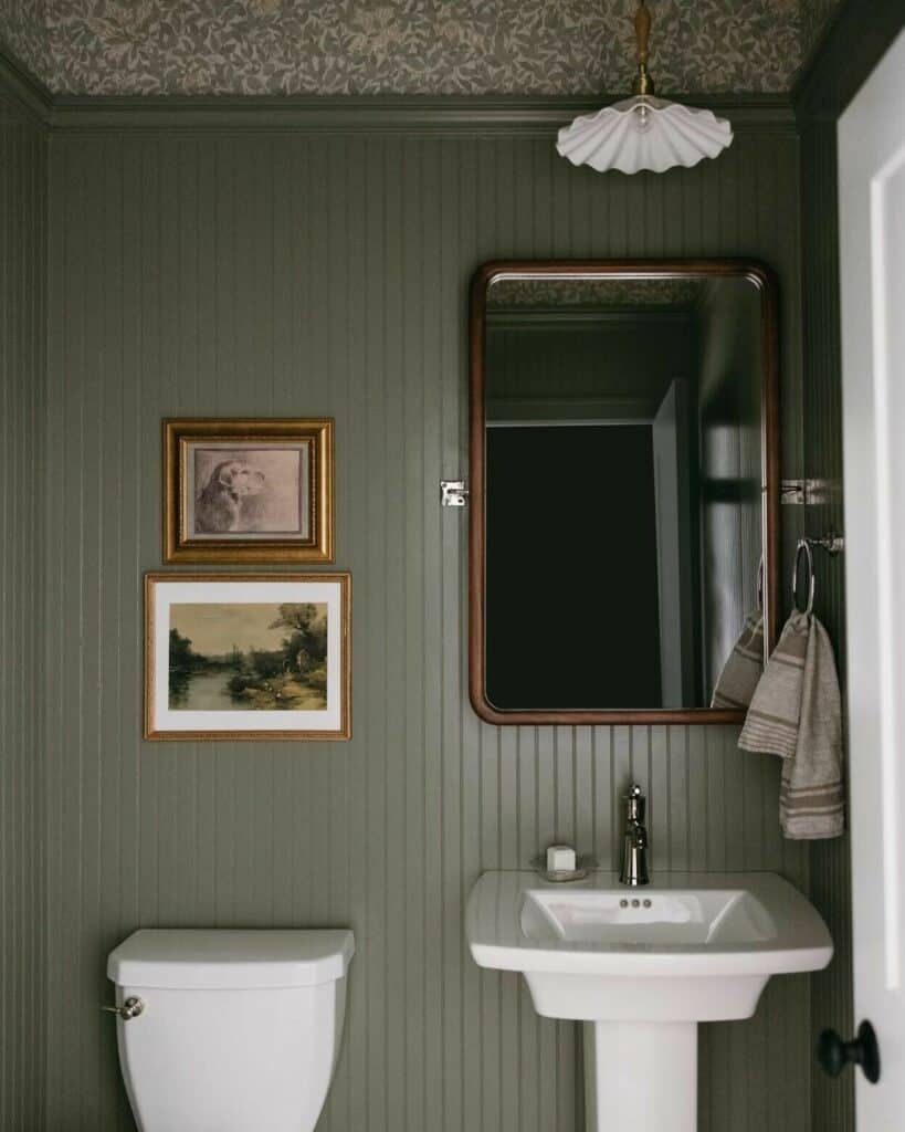 Small green cloakroom with Farrow and Ball Treron tongue and groove panelling, pedestal basin, gilt mirror and gallery wall with patterned wallpaper ceiling