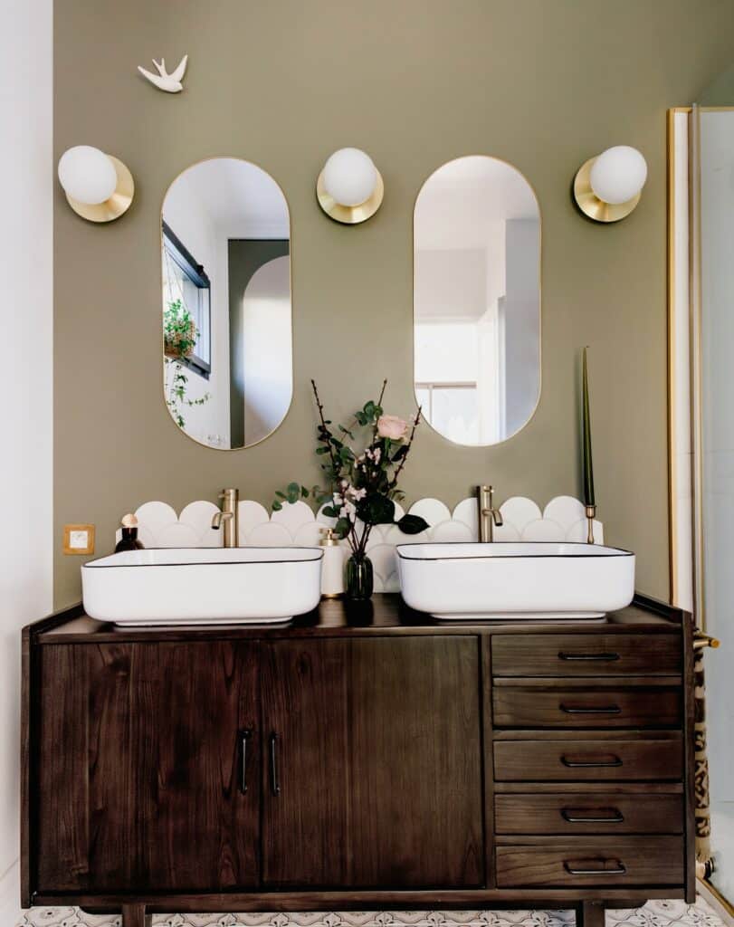 Sage green bathroom with Farrow and Ball Treron walls, dark wood double vanity, scallop edge splashback, twin oval mirrors and brass globe wall lights