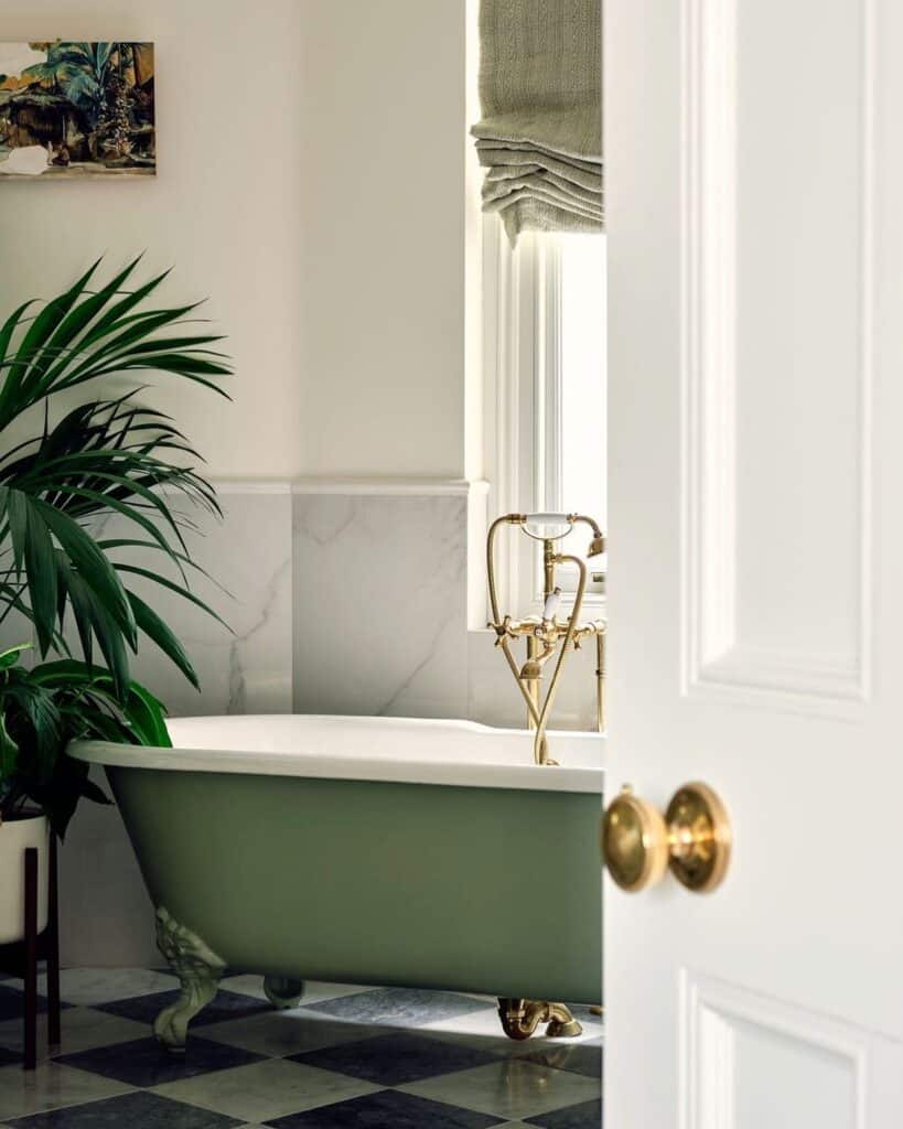 Sage green clawfoot bath painted in Farrow and Ball Calke Green with brass freestanding taps, marble half-panelled walls and a large indoor palm in a classic British bathroom