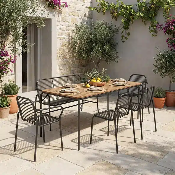 Normann Copenhagen Vig patio dining set with teak table, wire steel chairs and bench in a walled Mediterranean-style courtyard garden with olive trees and climbing plants.​​​​​​​​​​​​​​​​
