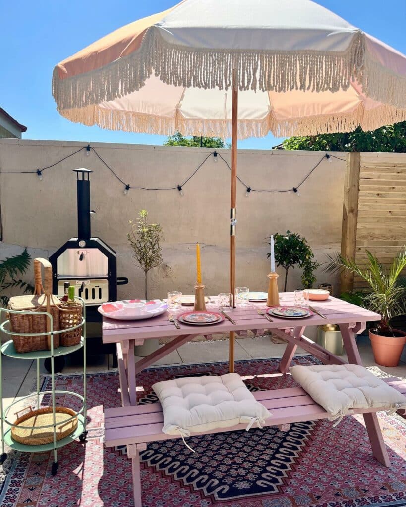 Walled courtyard garden painted Farrow & Ball Cinder Rose with pink picnic table, fringed parasol, pizza oven and string lights, a colourful small courtyard garden idea.​​​​​​​​​​​​​​​​