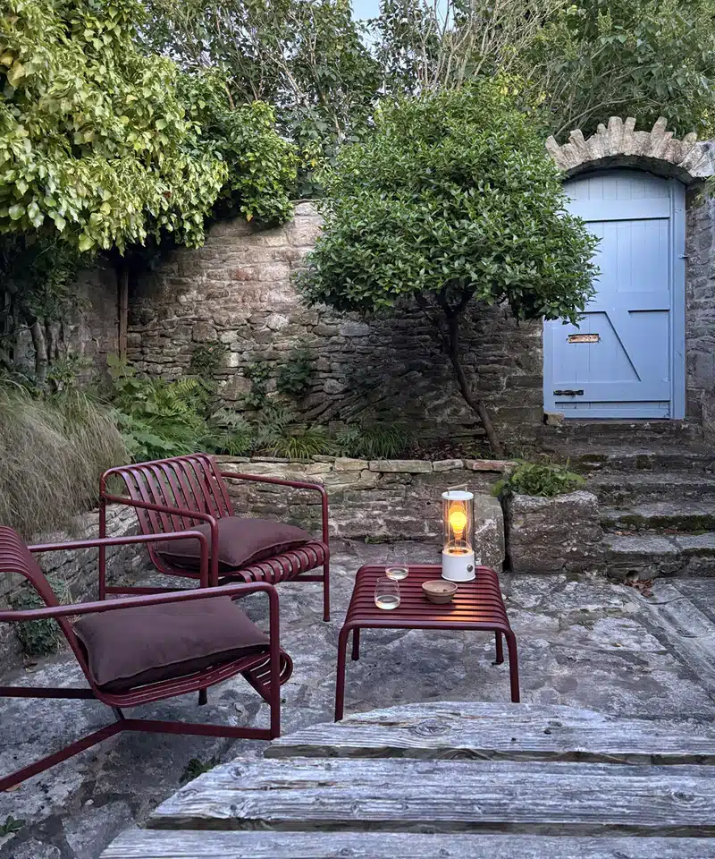 HAY Palissade Bordeaux lounge chairs and footstool with portable lamp at dusk in a walled courtyard garden with stone steps and blue painted door, Dorset.
