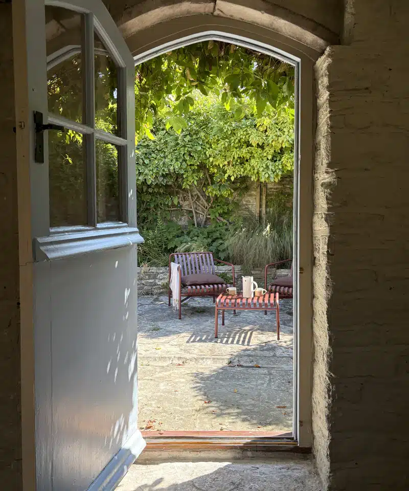 Walled courtyard garden with HAY Palissade lounge chairs on stone paving viewed through an arched stone doorway at a historic Dorset cottage.