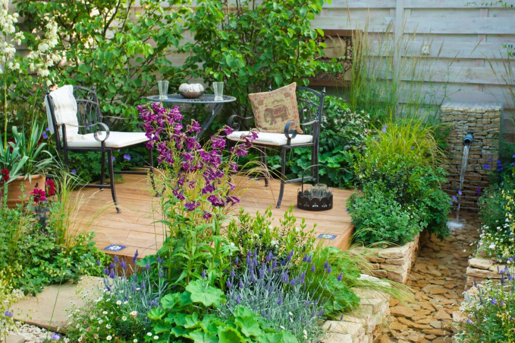 Lush walled courtyard garden with densely planted cottage-style borders, flagstone paving, wrought iron bistro chairs and a small water feature.​​​​​​​​​​​​​​​​