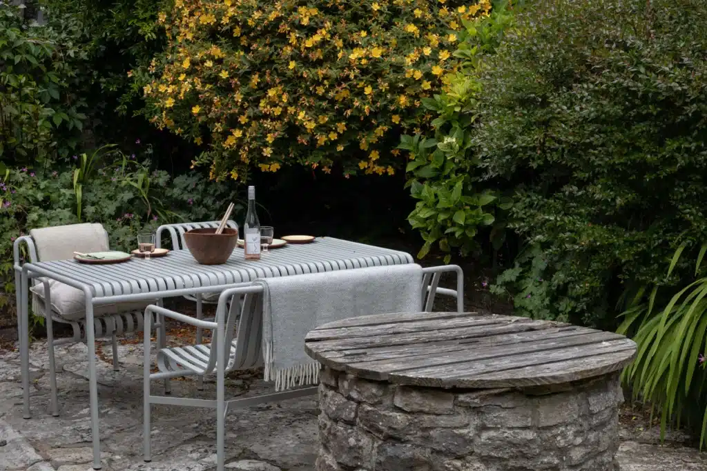 HAY Palissade Sky Grey outdoor dining table set in a lush courtyard garden with dense yellow flowering shrubs and established planting, Purbeck Cottage Dorset.​​​​​​​​​​​​​​​​
