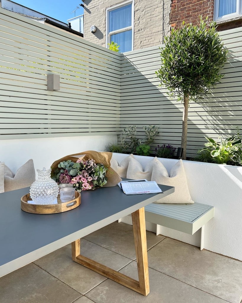 Modern courtyard garden with Farrow & Ball Mizzle slatted fence panels, built-in bench seating and olive tree standard, a contemporary small courtyard garden idea.​​​​​​​​​​​​​​​​