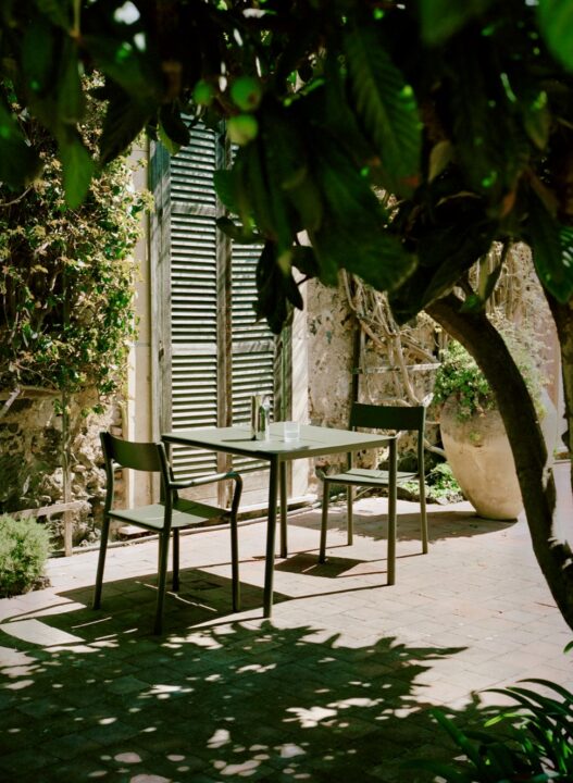 Compact green metal outdoor table and chairs set in a shaded small courtyard garden, surrounded by lush greenery and rustic stone walls. A charming example of courtyard garden ideas for intimate outdoor dining.