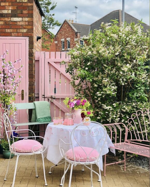 Charming vintage courtyard garden with a white outdoor dining set, pink cushions, and a pastel pink painted gate. A romantic and stylish inspiration for small courtyard garden ideas.
