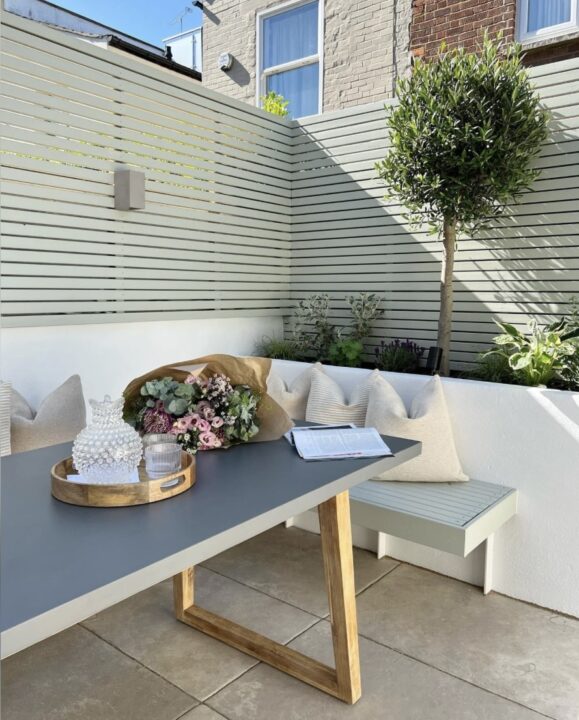 Modern courtyard garden with built-in planter boxes, a sage green slatted wall, and a cosy seating area with a sleek dining table. A stylish example of courtyard garden ideas for small outdoor spaces.