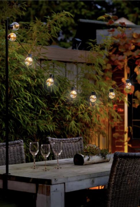 Courtyard garden with festoon string lights draped over a wooden outdoor dining table, creating a warm and inviting atmosphere for evening gatherings. A cosy lighting idea for small courtyard garden ideas