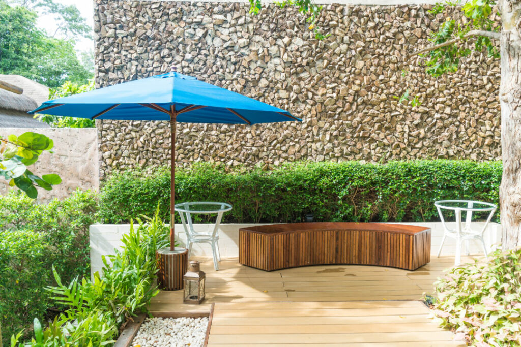 Modern courtyard garden with a wooden deck, curved bench seating, a blue patio umbrella, and lush greenery against a textured stone wall. A stylish and functional example of courtyard garden ideas for outdoor relaxation.
