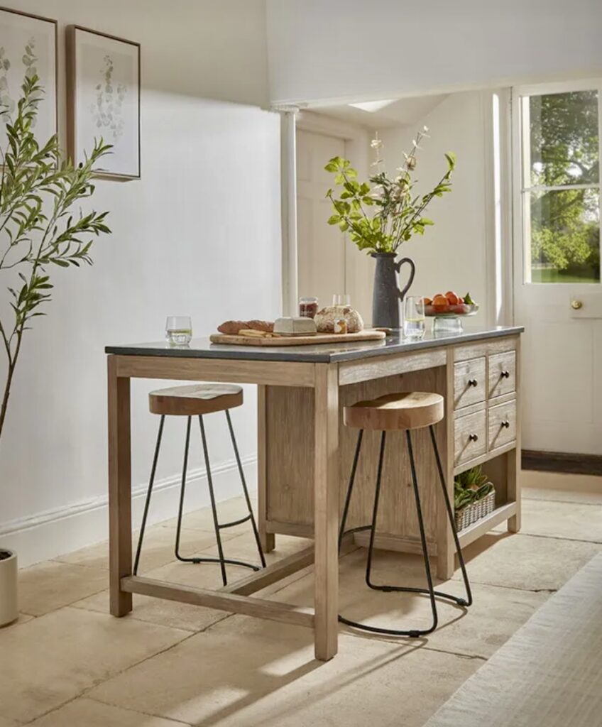 A freestanding kitchen breakfast bar featuring the Maria Kitchen Island Unit with a solid wooden frame, natural wood grain finish, and a contrasting dark worktop. Paired with stylish wood and metal bar stools, this rustic yet functional island adds warmth and elegance to any kitchen.