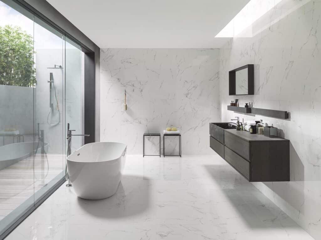 White marble bathroom with freestanding bath, dark floating double vanity, glass shower enclosure and floor-to-ceiling Carrara marble tiles