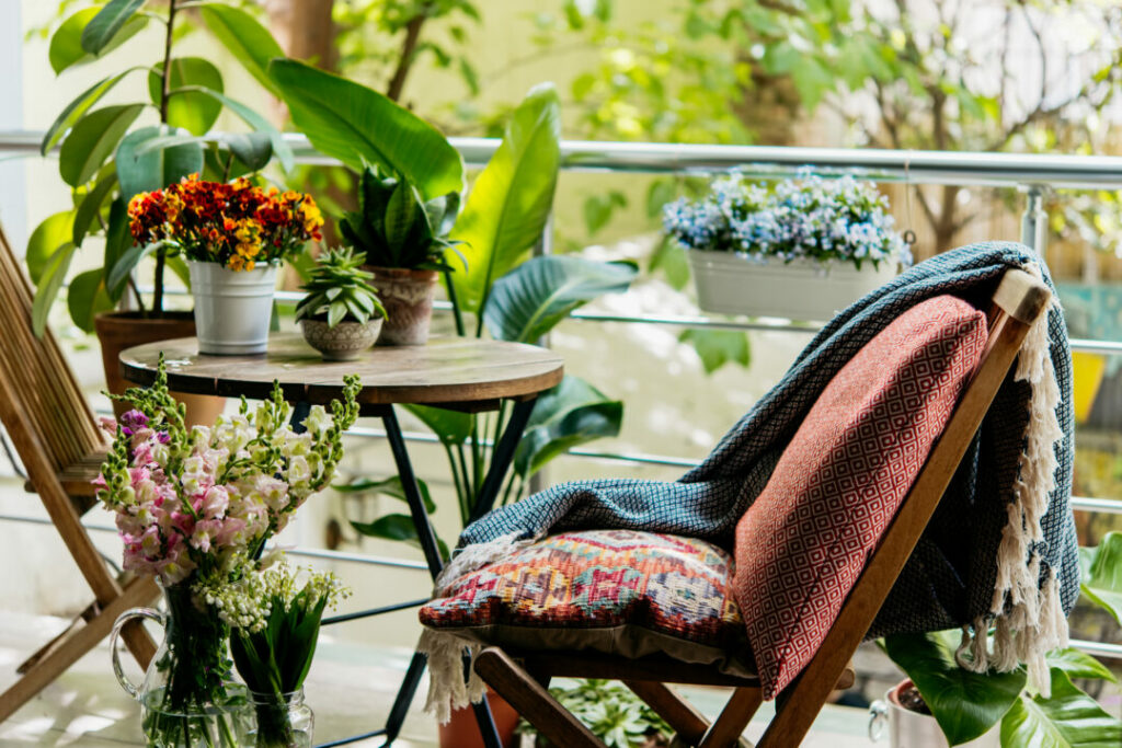 Cosy small balcony with railing planters, potted plants, and a wooden chair with cushions and throws – a relaxing small balcony idea.