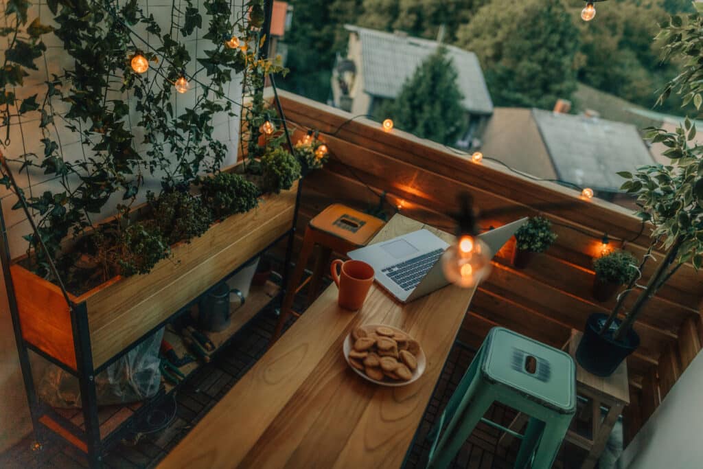 Small balcony with slim wooden bar table, stools, string lights and railing planter creating a cosy work-from-home seating area