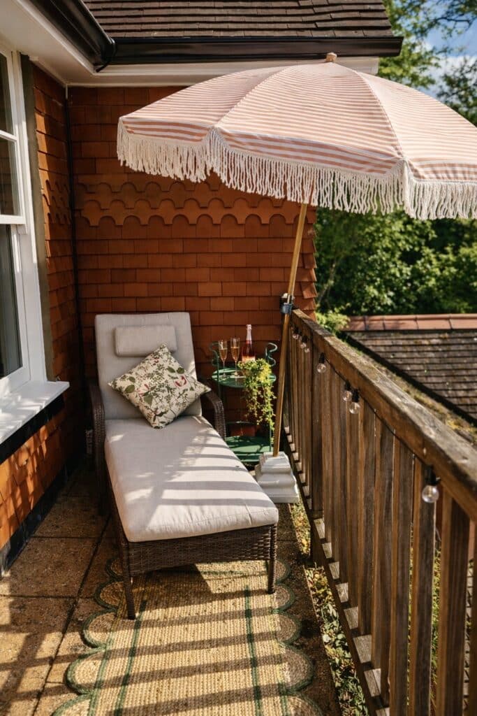 A narrow Victorian balcony styled with a wicker sun lounger, fringed striped parasol, drinks trolley and scalloped jute runner — small balcony ideas that transform a compact outdoor space.