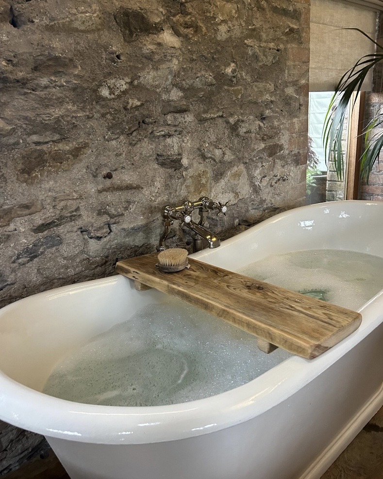 Reclaimed wood bath caddy across a white freestanding bath with brass taps against a raw stone wall