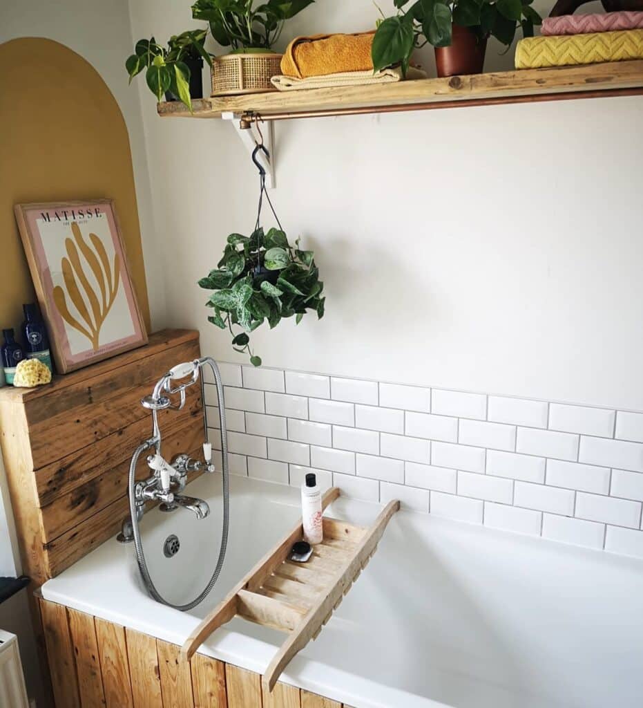 Rustic bathroom with Farrow and Ball Sudbury Yellow painted wall, reclaimed wood bath panel, white metro tiles and wooden bath rack with plants