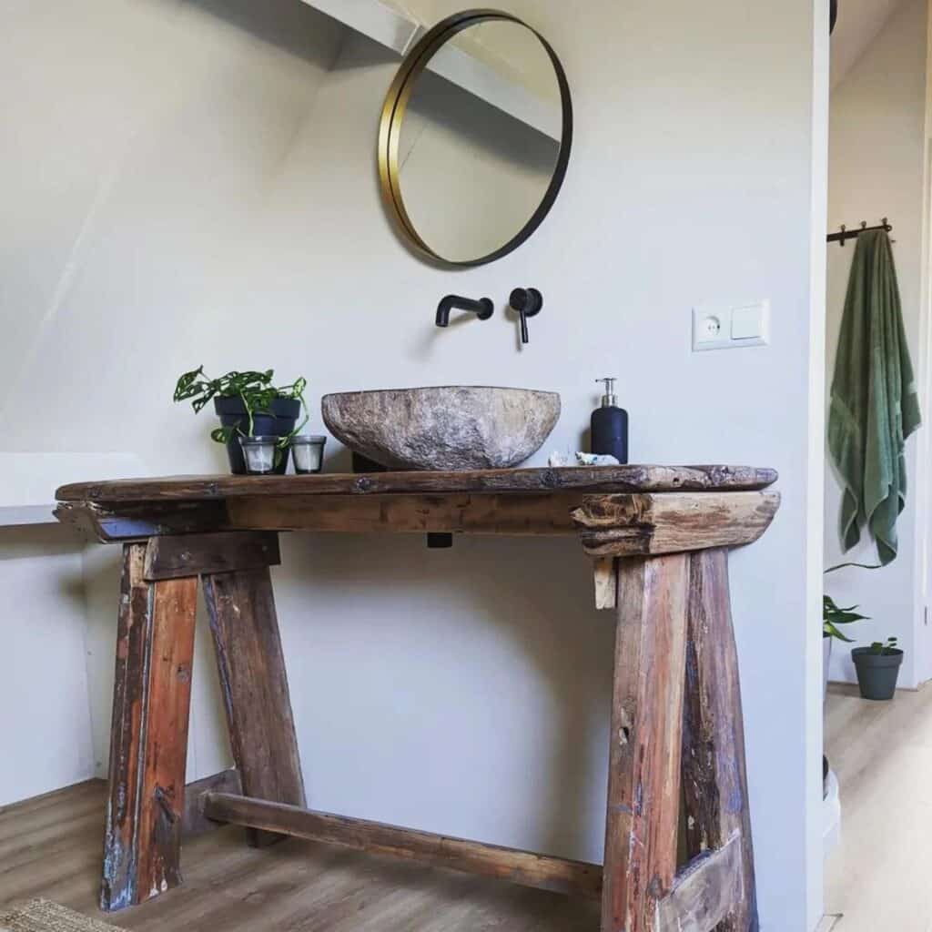 River stone vessel basin on a reclaimed wood trestle vanity with black wall-mounted tap and round brass mirror under a sloped ceiling
