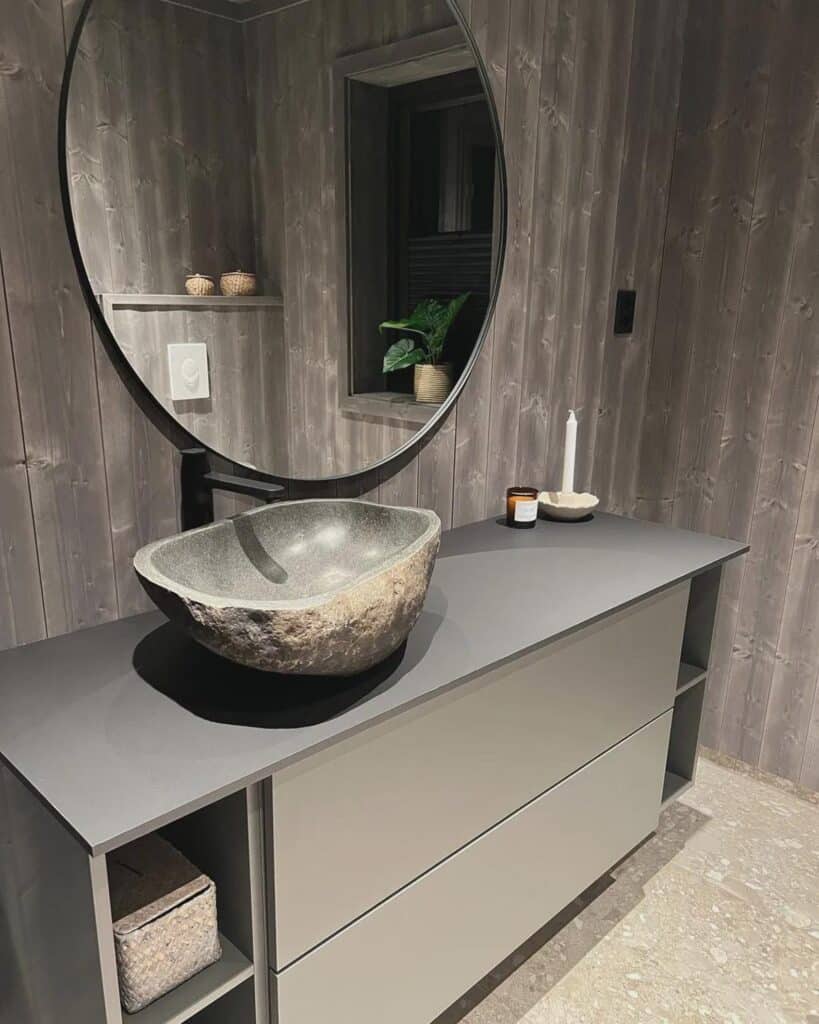 River stone vessel basin on a dark grey modern vanity against a grey wood-panelled wall with round black mirror