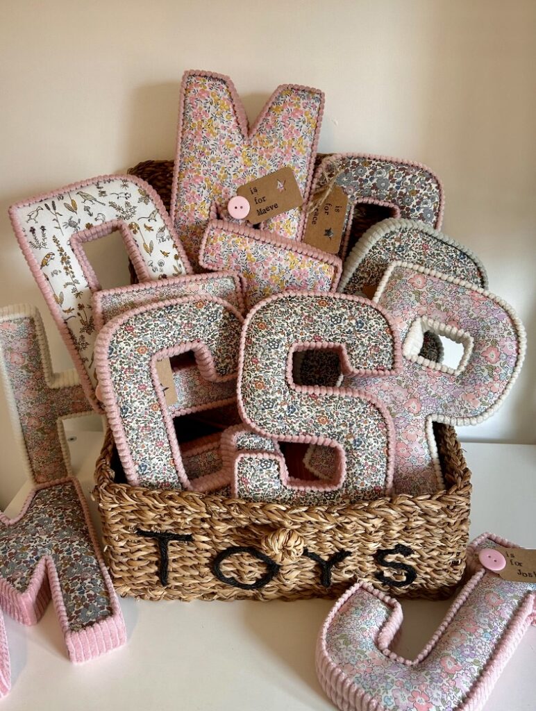 Handmade Liberty print nursery fabric letters by Buttons and Bells, arranged in a woven basket with various initials in soft pastel floral patterns.