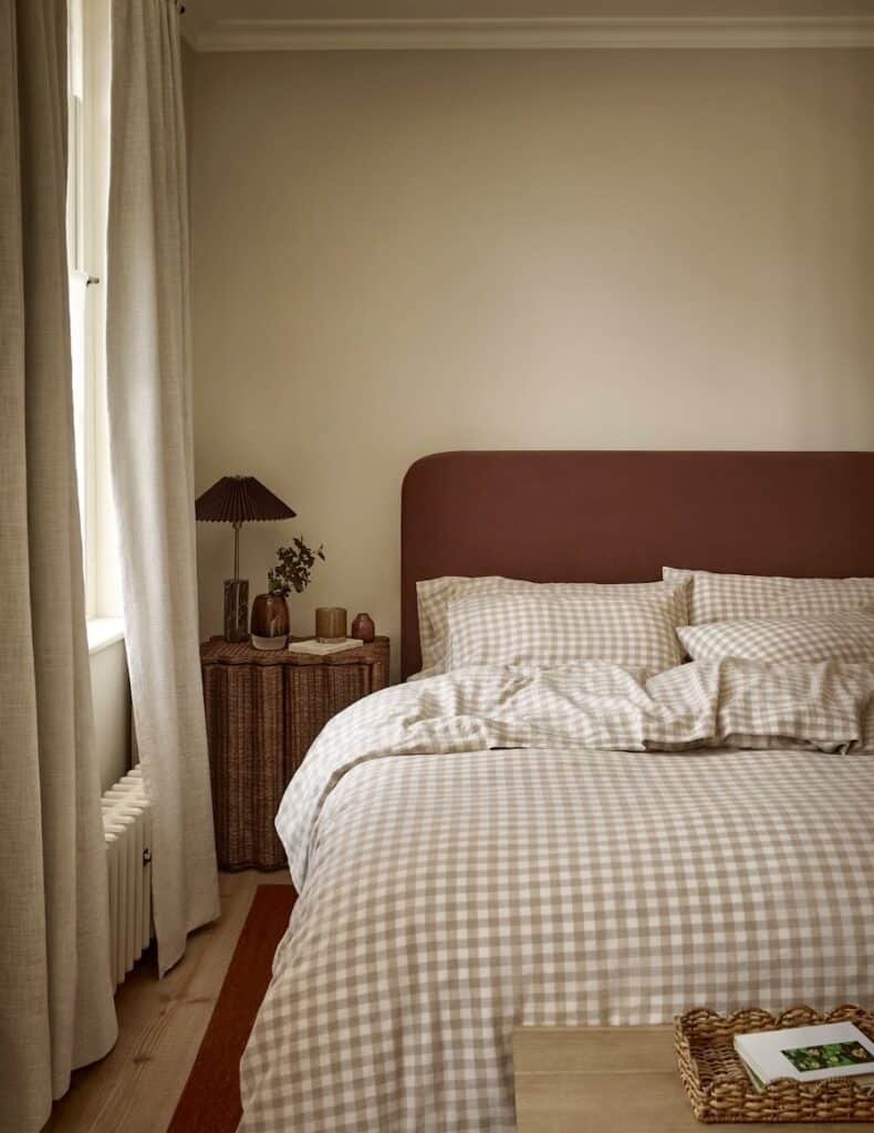Warm neutral cottage bedroom with oatmeal gingham linen bedding, a rust upholstered headboard, a scalloped rattan bedside table and a pleated lampshade
