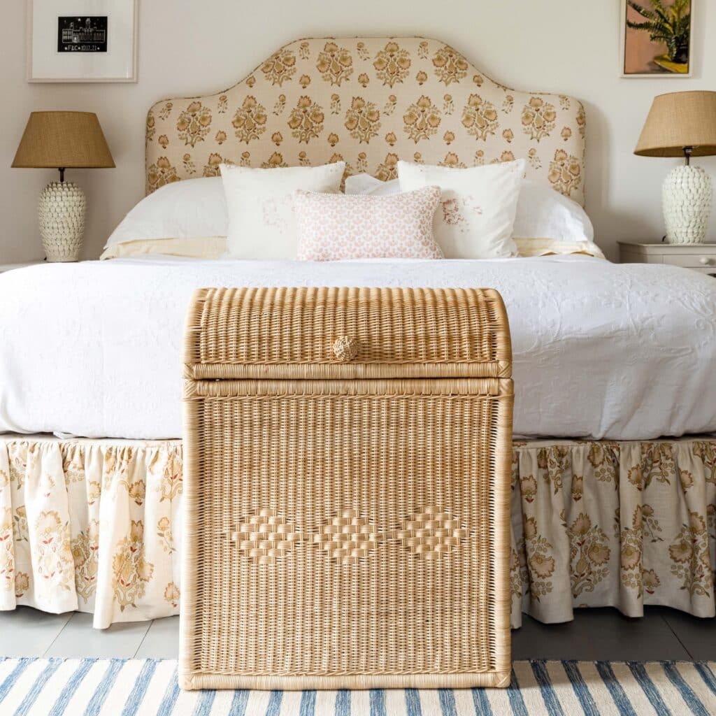 Warm neutral cottage bedroom with a handwoven rattan chest at the foot of the bed, a block-print scalloped headboard, a floral gathered valance and woven lampshades