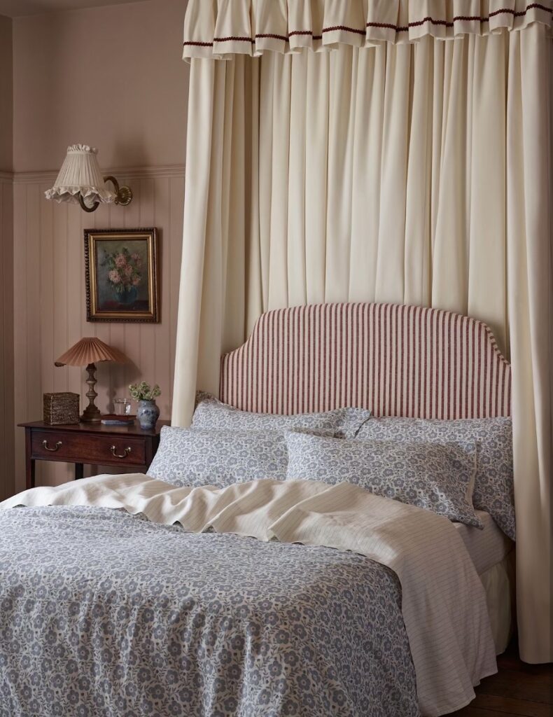 Cottage bedroom with a ceiling-hung cream fabric canopy and ric-rac pelmet, a red striped headboard and Folk Floral blue cornflower linen bedding