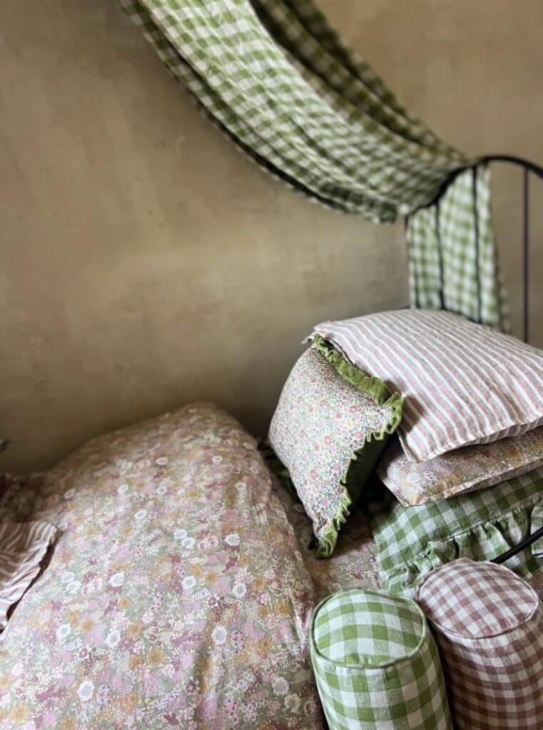 Layered bedding in cottage bedroom with Liberty floral quilt, green gingham canopy and mixed patterned cushions for a warm, cosy look.