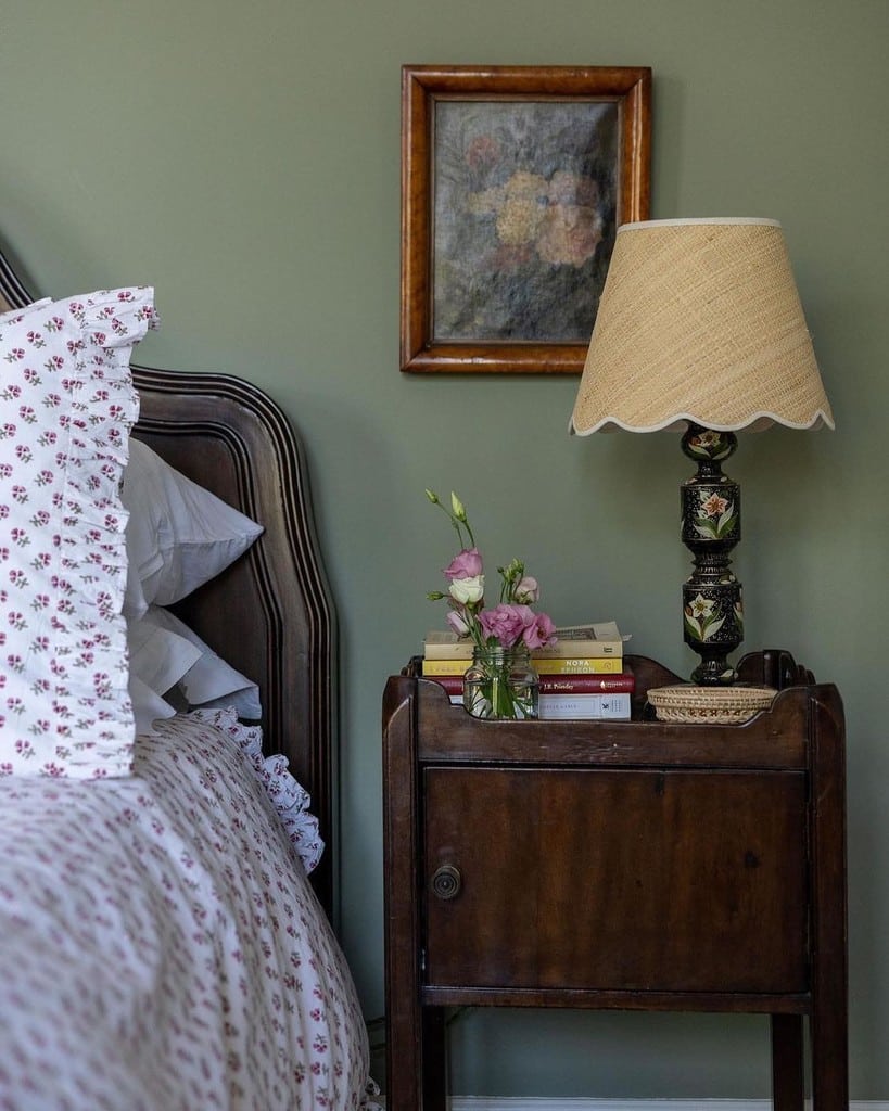 Cottage bedside with a scalloped raffia lampshade on an antique painted lamp base, a ruffled floral pillowcase and pink roses against Lichen green walls