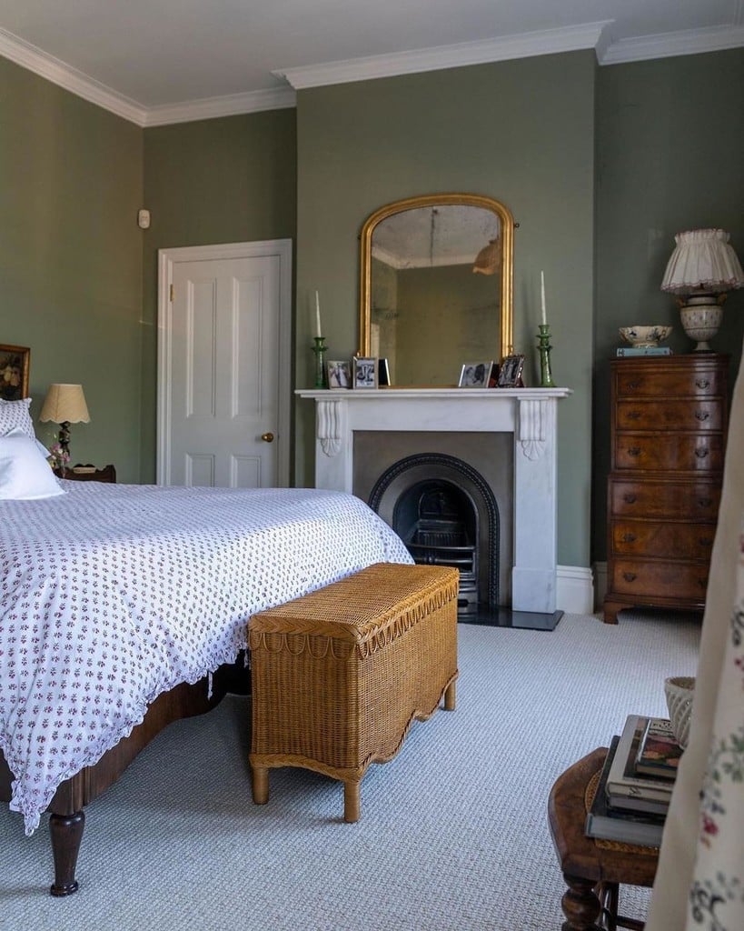 Sage green cottage bedroom painted in Farrow & Ball Lichen, with a block-print floral bedspread, a scalloped wicker trunk, a marble fireplace and a gilt mirror