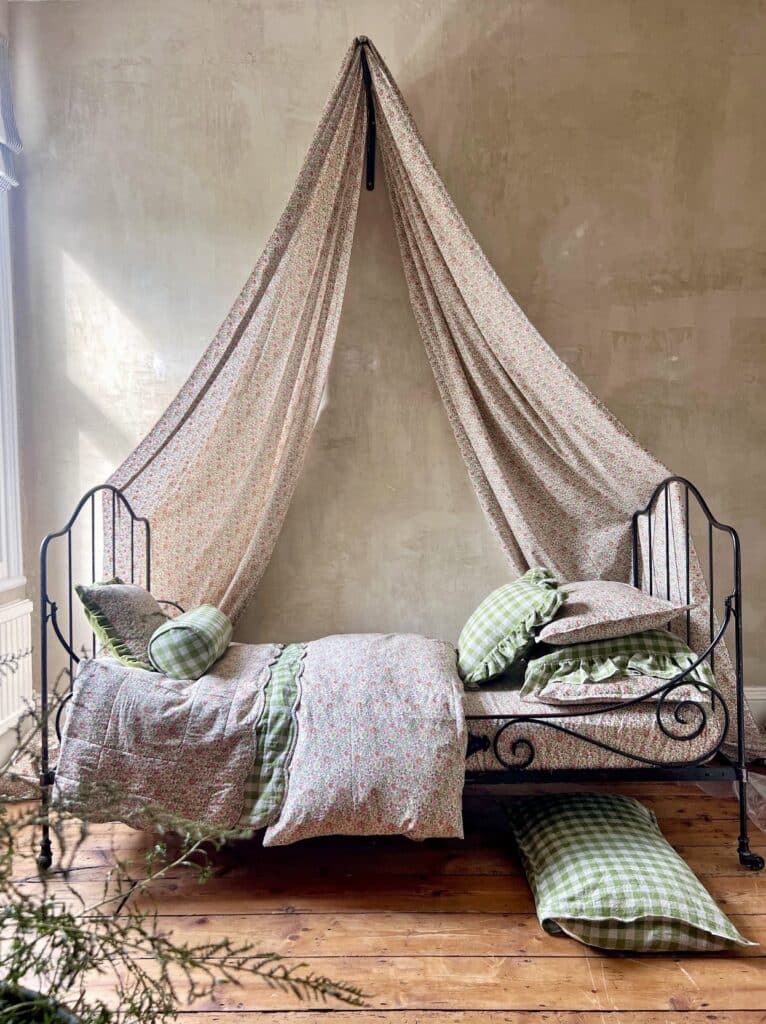Cottage bedroom idea with Liberty floral fabric draped as a canopy over an iron daybed, layered with floral bedding and green gingham ruffle pillows