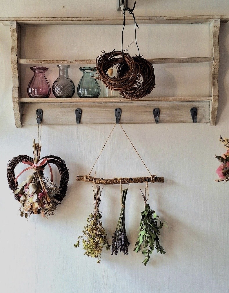 Cottage wall vignette with a rustic peg shelf, coloured glass bud vases, twig wreaths, a dried flower heart and a hanging bar of dried lavender, oregano and eucalyptus