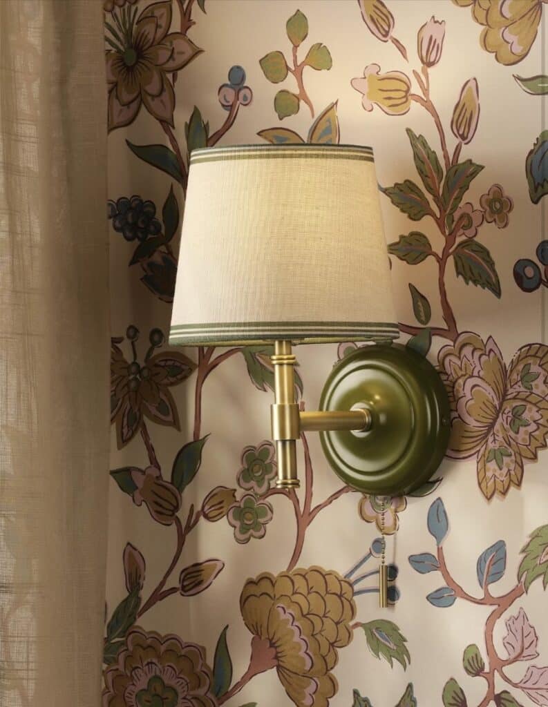 Joules Upton wall light with a green painted mount, antique brass arm and striped linen shade against floral wallpaper in a cottage bedroom