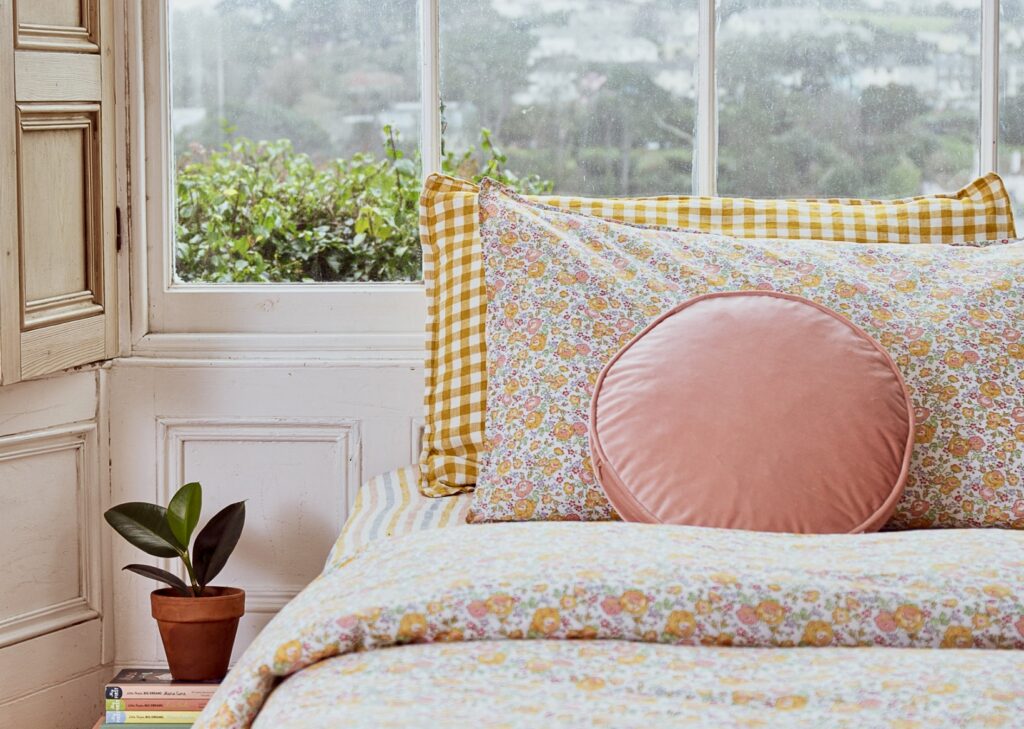 A cosy cottage bedroom featuring a floral bedding set by Poppy and Honesty, made with Liberty fabric. The bedding is styled with gingham pillows in yellow and a round pink cushion, adding layers of colour and texture. A terracotta plant pot sits on a windowsill, completing the charming, nature-inspired design.