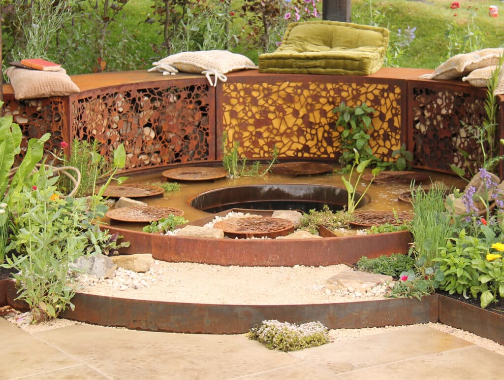 Gravel garden ideas featuring a corten steel sunken seating area with gravel base, circular fire pit and planted borders.