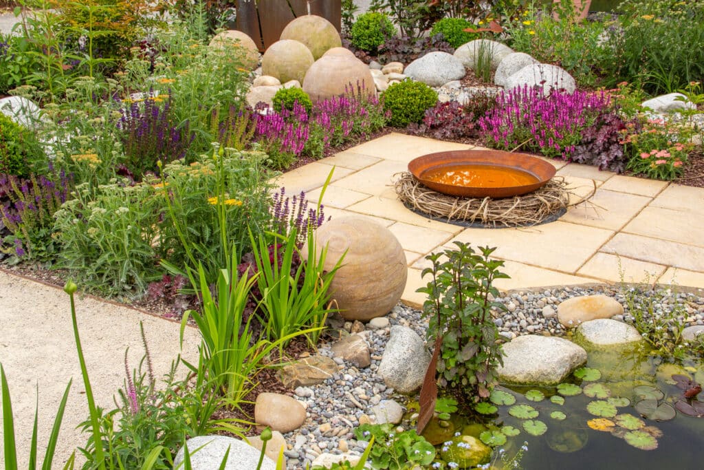 Gravel garden ideas with a pond edge, boulders and colourful planting beside a small seating area