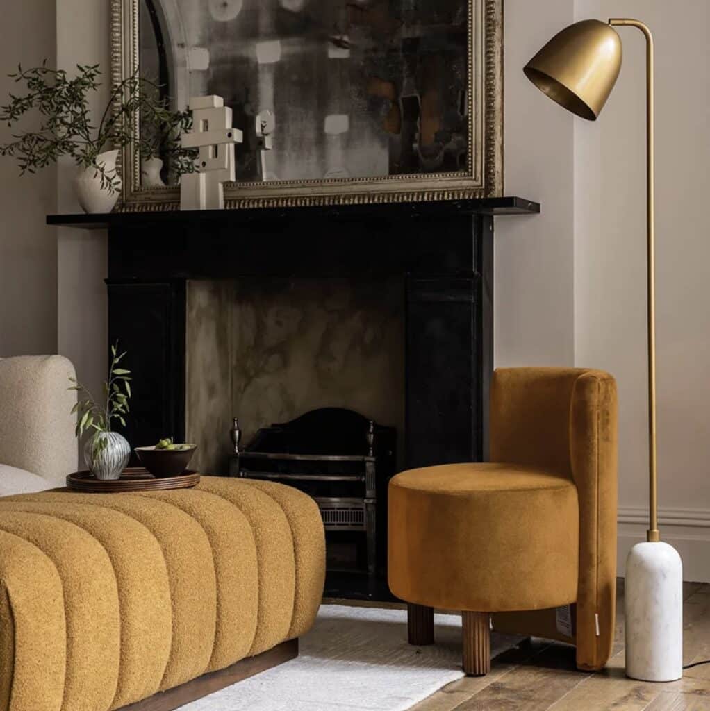 A sophisticated living space featuring a mustard colour velvet occasional chair alongside a matching ottoman. The mustard colour furniture contrasts beautifully with the black fireplace, creating a warm and elegant aesthetic. A brass floor lamp complements the mustard colour tones, enhancing the cosy yet refined ambiance.