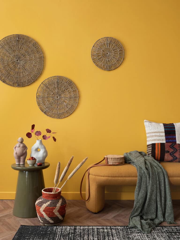 Mustard-colour wall with woven rattan wall baskets, a low mustard bench, olive throw and patterned cushion in a relaxed living room corner.