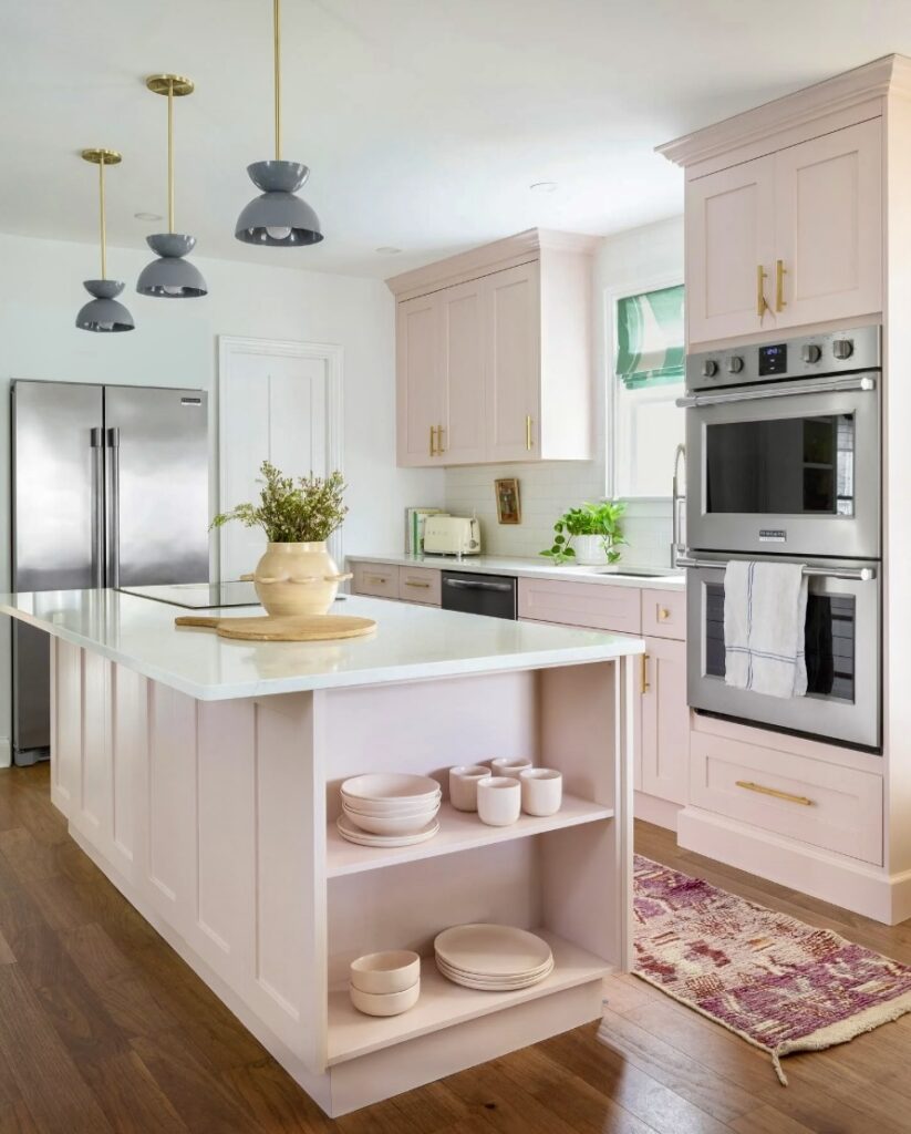 Soft pink kitchen featuring Calamine cabinetry by Farrow & Ball, a white freestanding kitchen island with open shelving, gold hardware, and muted green accents. The space is complemented by white countertops, stainless steel appliances, and natural wood flooring, creating a charming cottage-style aesthetic.