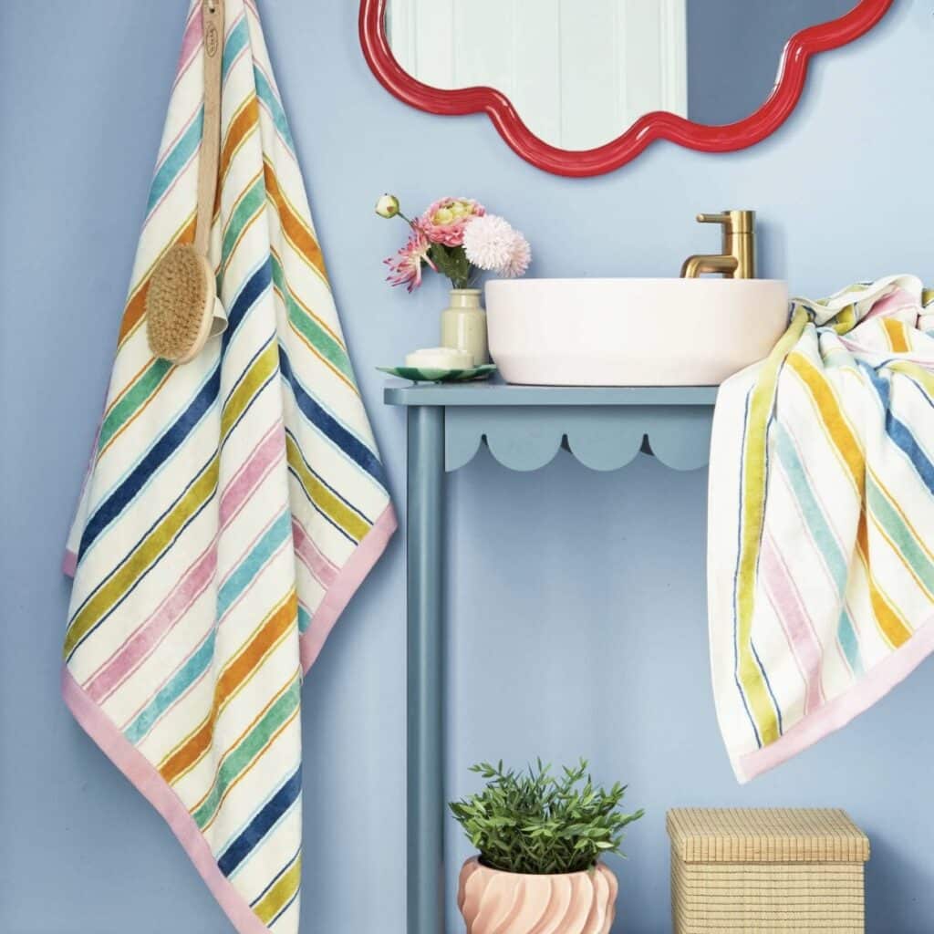 Rainbow stripe towel hanging beside a blue vanity with scalloped trim and red wavy mirror in a small bathroom
