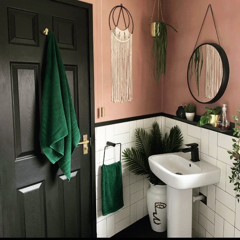 Small bathroom with Sulking Room Pink walls above white metro tiles, black round mirror, green towels and trailing plants — budget small bathroom ideas UK