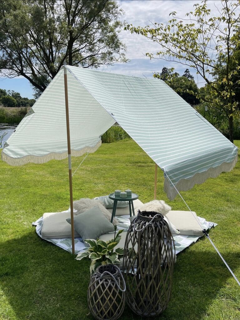 Sage green striped garden canopy tent with tassel fringe on a lawn with floor cushions and rattan lanterns, DIY garden shade ideas