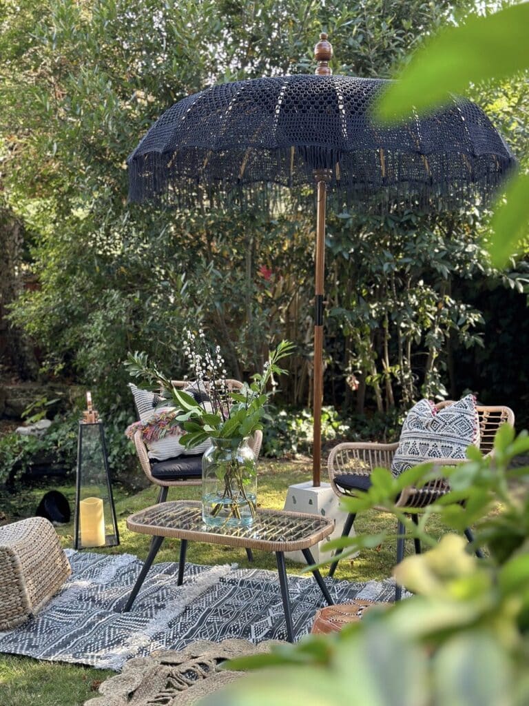 Black macramé boho garden parasol over rattan chairs and woven rug on a lawn with lush green planting