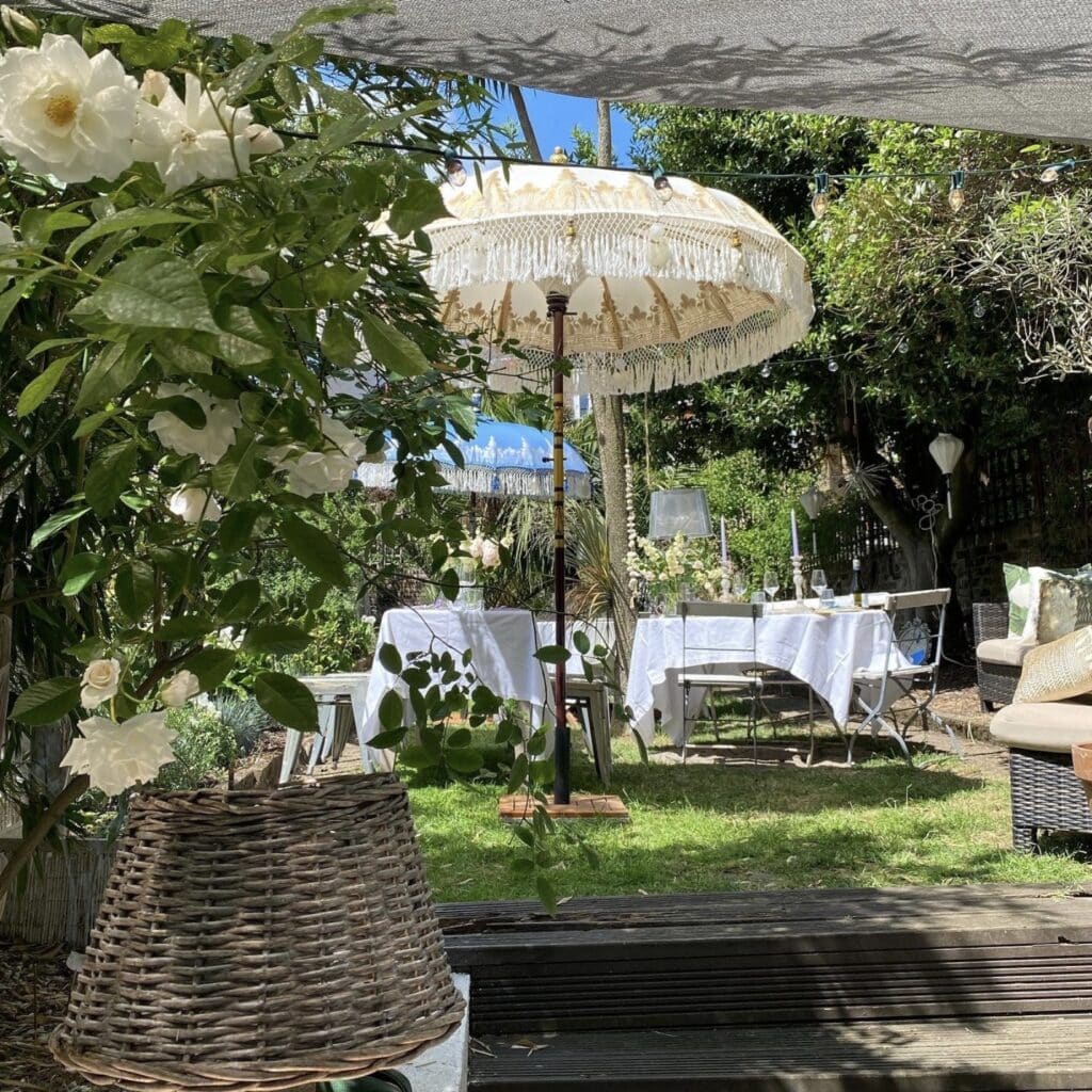 Hand painted Balinese cream and gold garden parasol with macramé fringe over an outdoor dining table with white roses and festoon lights