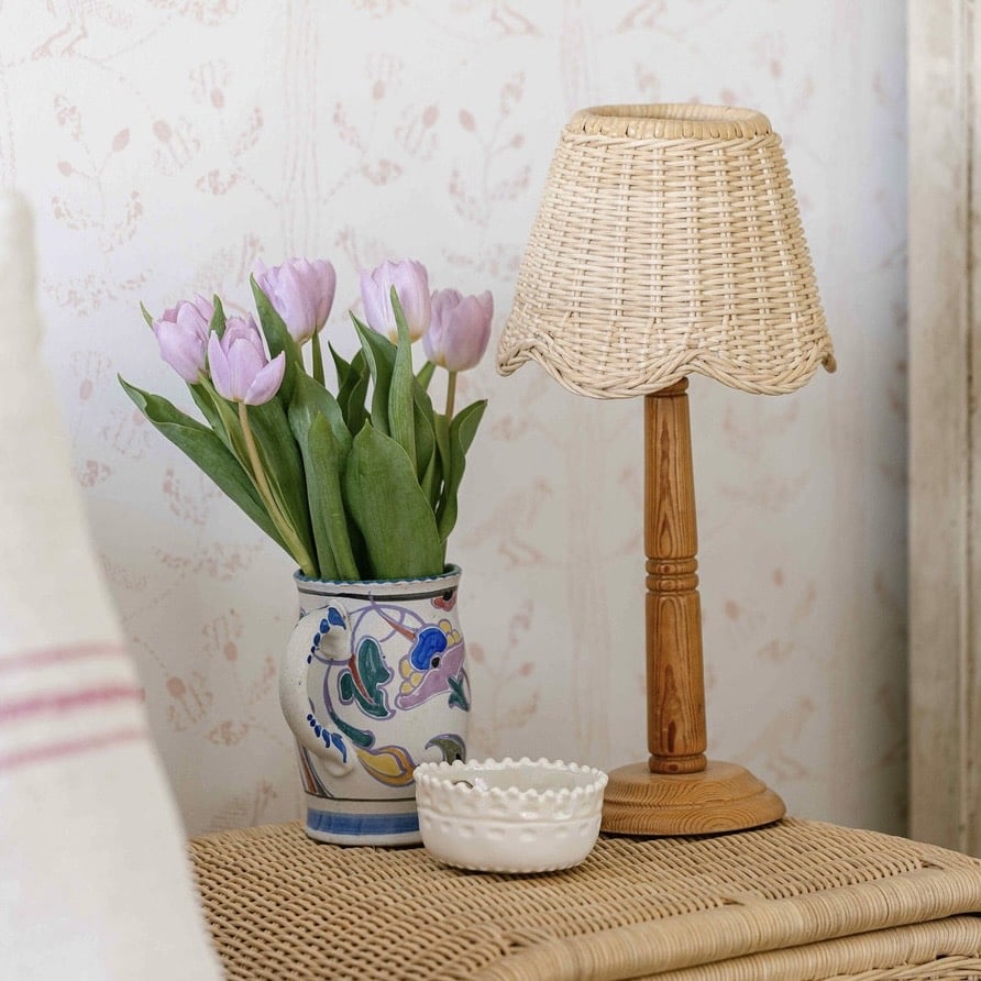 Rattan scalloped lampshade on a wooden table lamp beside a vase of tulips on a woven bedside table.