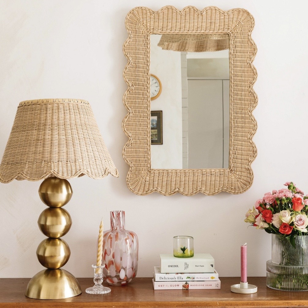 Rattan scalloped edge mirror above a wooden sideboard with a brass lamp, candles and flowers for relaxed living room styling.